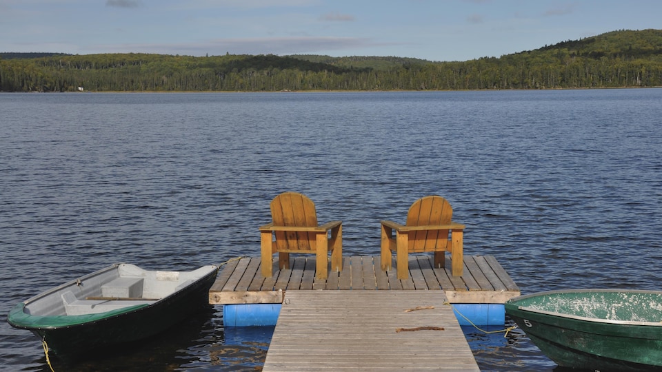 Deux chaises de style Adirondack se trouvent sur le quai d’un lac. Deux chaloupes flottent de chaque côté.