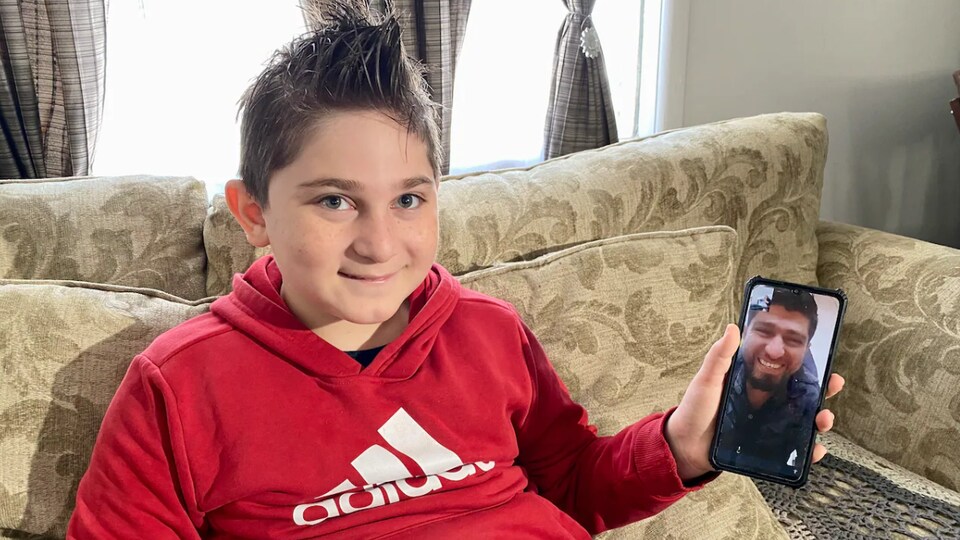 Adnan Kharsa, 9 ans, est assis sur un canapé et tient un téléphone cellulaire dans sa main gauche. 