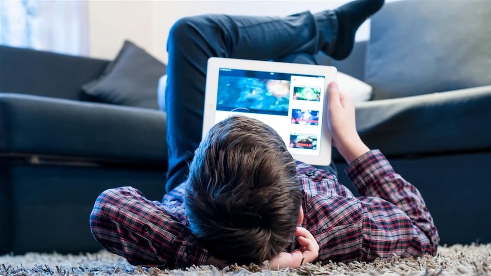 Un adolescent regarde une tablette, allongé sur le plancher du salon.