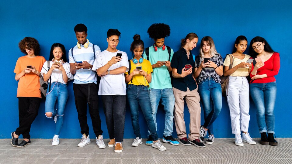 Un groupe de jeunes personnes consultent leur téléphone intelligent debout le long d'un mur bleu.
