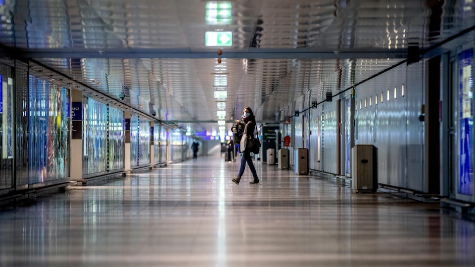 Un long couloir de l'aéroport de Francfort, en Allemagne, est pratiquement désert. On voit au loin quelques voyageurs, dont une dame masquée qui marche vers la gauche. 