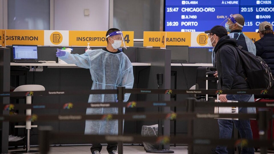 Des voyageurs font la file près d'un employé de l'aéroport portant un masque, une visière et une tenue protectrice.