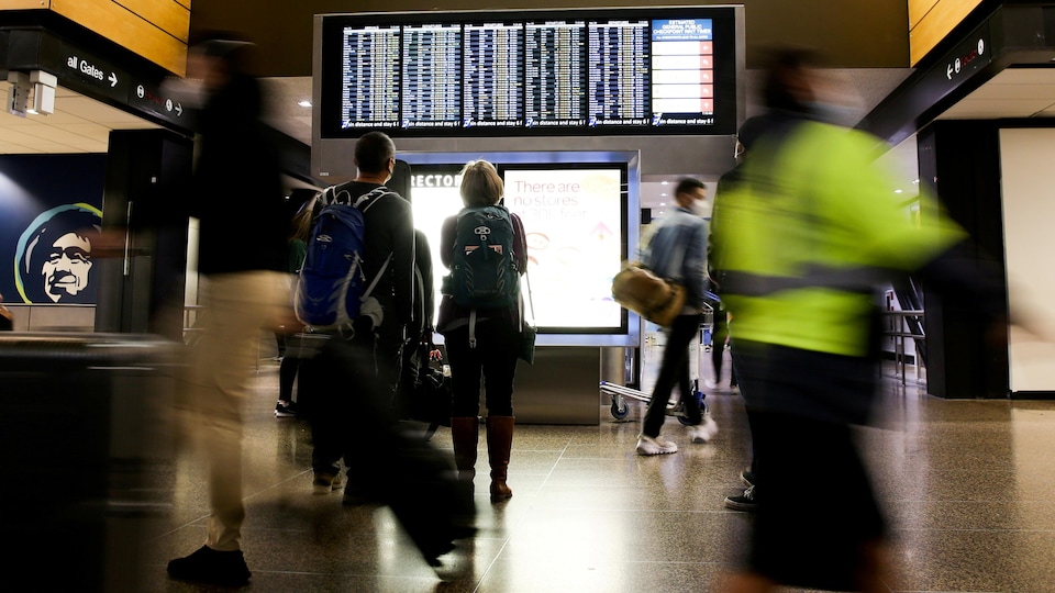 Des personnes regardent l'horaire de départ des vols sur un écran dans un aéroport.