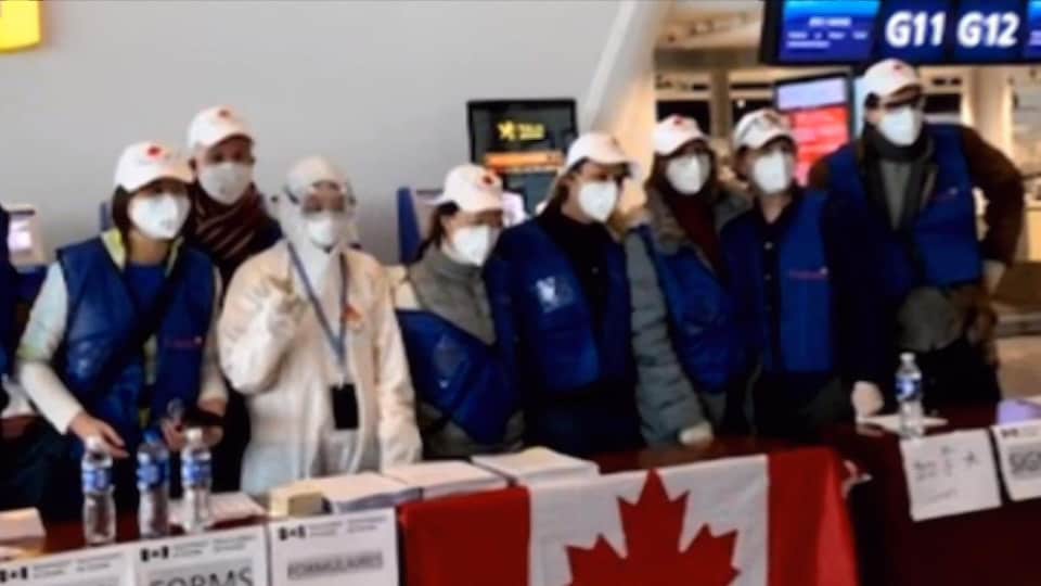 Plusieurs personnes masquées sont debout derrière une table devant laquelle a été installé un drapeau canadien. 