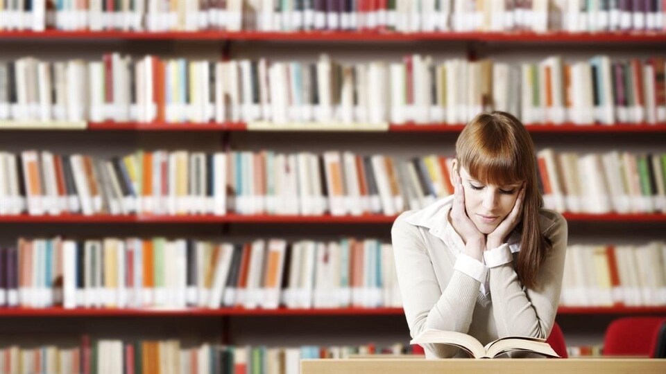 Une femme lit dans une bibliohtèque. Des étagères remplies de livres sont derrière elle