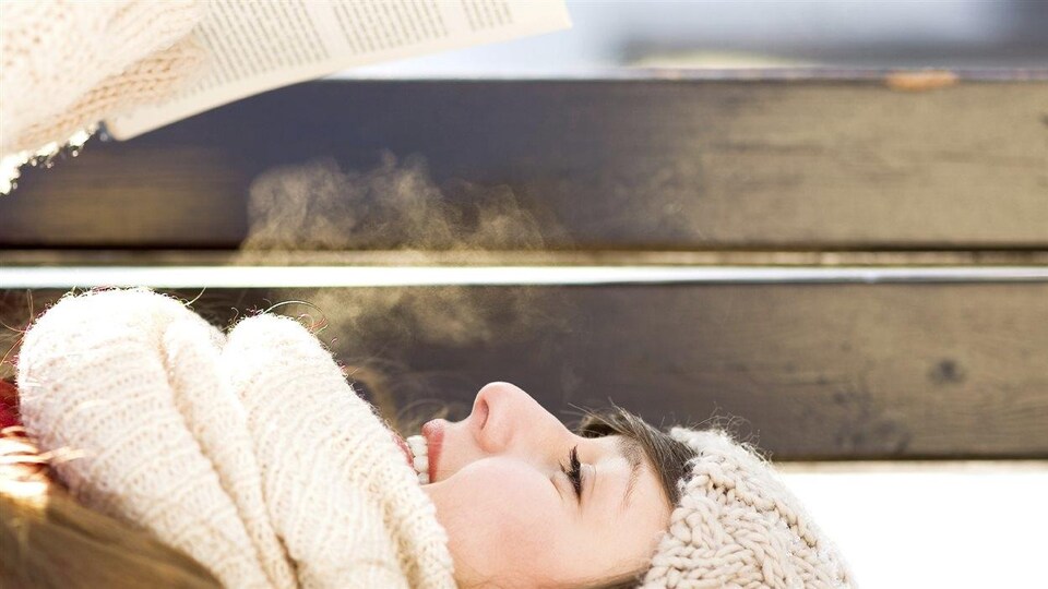 Femme lisant couchée sur un banc en hiver, à l'extérieur, avec un foulard, une tuque et des mitaines.