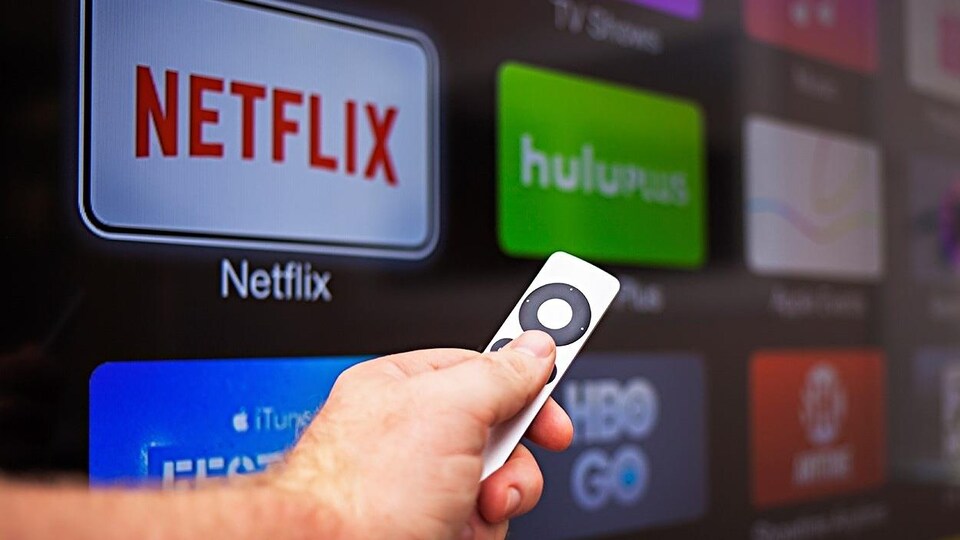 Une main tient une télécommande de télévision intelligente sur laquelle on voit les logos de plateformes de diffusion.