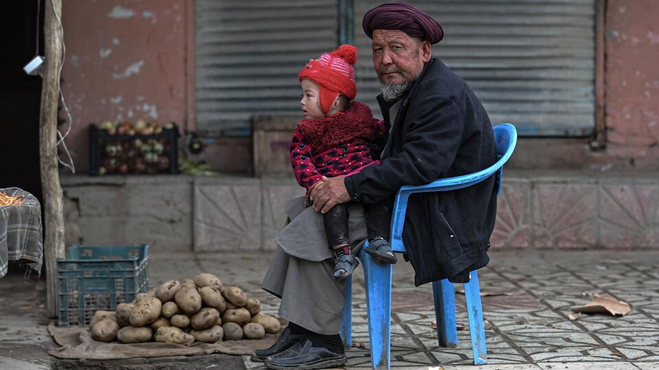 Un homme portant un enfant sur ses genoux devant quelques pommes de terre posées sur un trottoir.