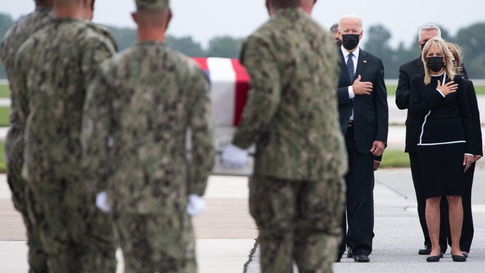 En arrière-plan, Joe Biden tient sa main sur le cœur. À l'avant-plan, des militaires portent un cercueil.