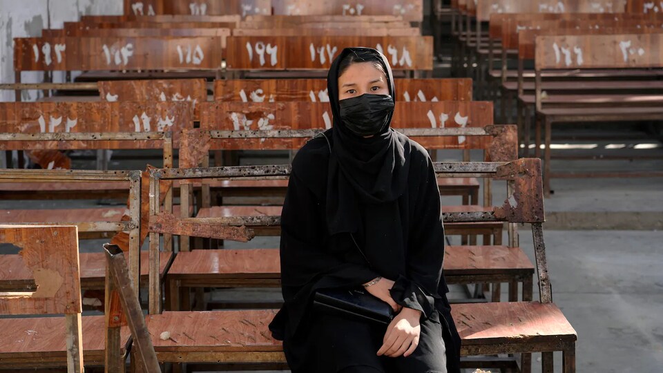 Une jeune Afghane vêtue d'une robe noire et d'un masque est assise sur un banc d'école.
