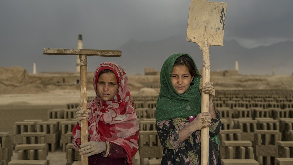 Deux jeunes filles tiennent une pelle et un pieu devant un terrain couvert de briques.