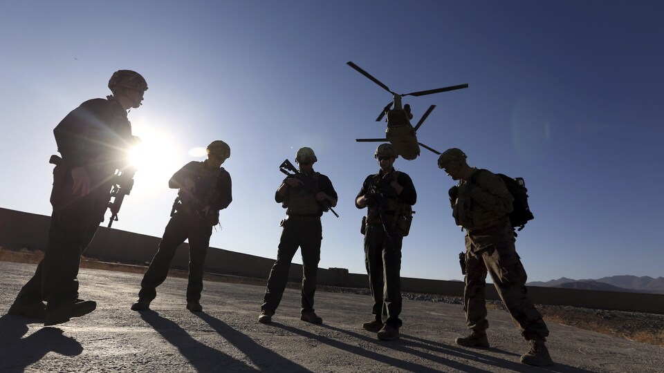 Cinq soldats américains attendent sur le tarmac pendant qu'un hélicoptère survole les lieux.