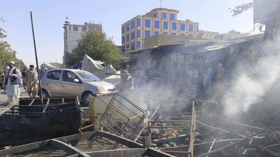 De la fumée s'échappe de divers objets sur le sol, avec en arrière-plan des Afghans qui se tiennent debout devant des magasins endommagés. 