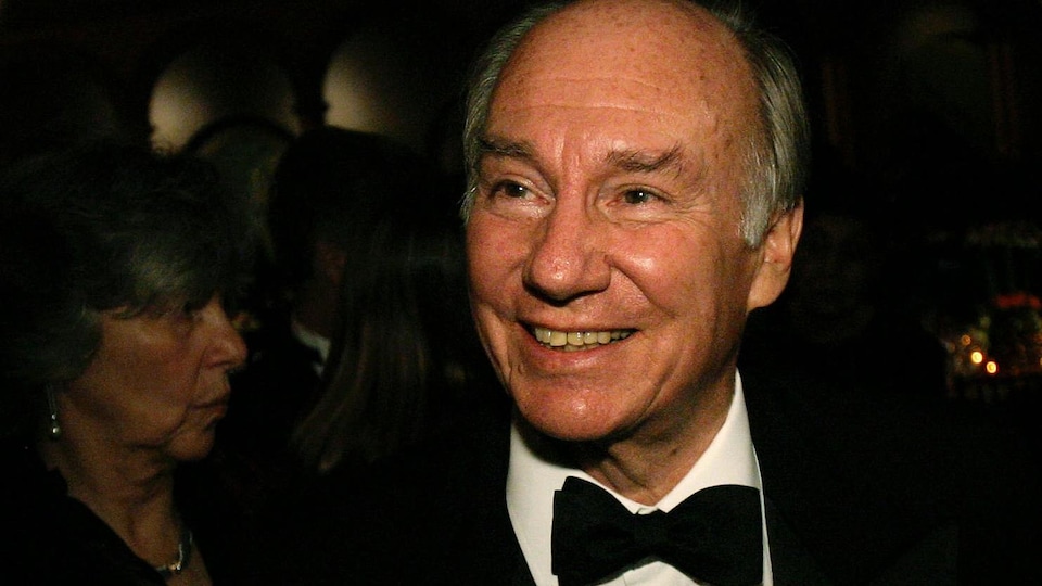 Le prince porte un complet noir et un noeud papillon. Il parle avec d'autres hommes.