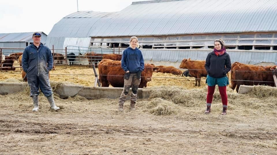 Un couple de producteurs agricoles et Émilise Lessard-Therrien sourient à la caméra devant un enclos de boeufs.