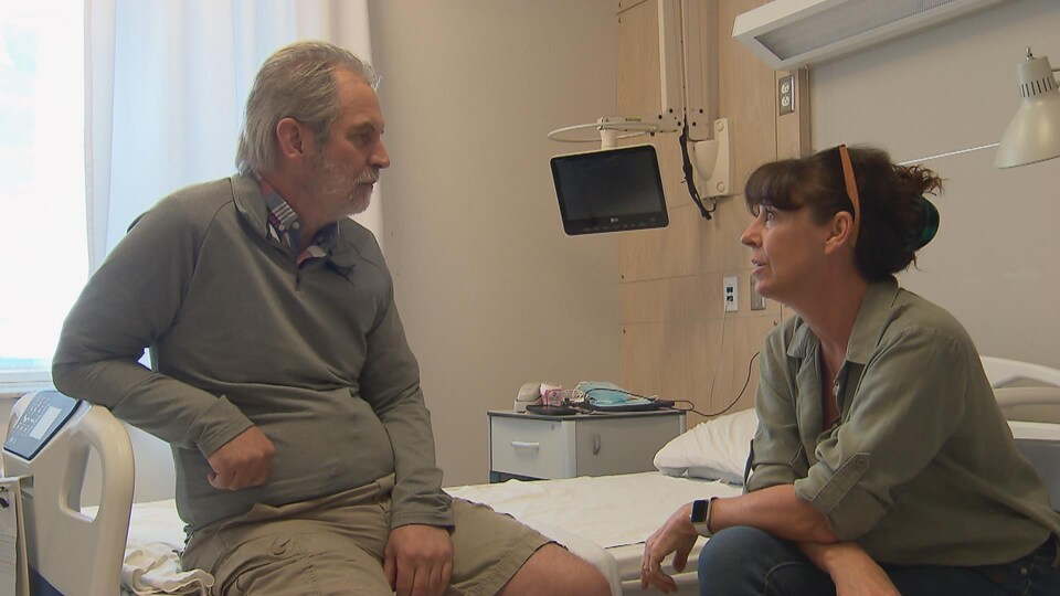 Un homme assis sur un lit d'hôpital et une femme assise en face de lui se regardent et discutent. La scène se passe dans une chambre d'hôpital.