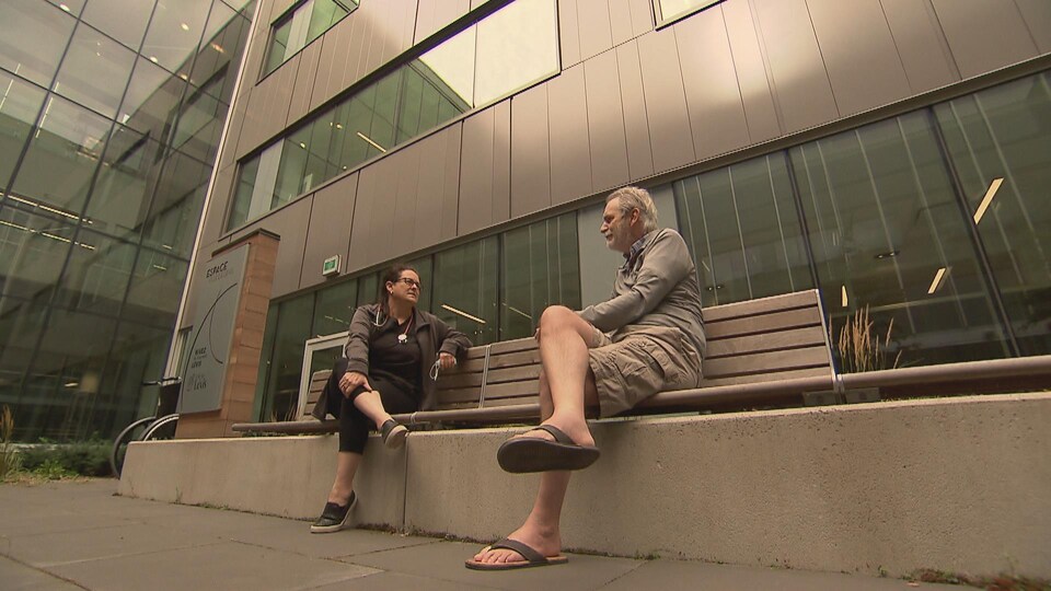 Une femme et un homme assis sur un bac à l'extérieur d'un grand bâtiment discutent en se regardant.