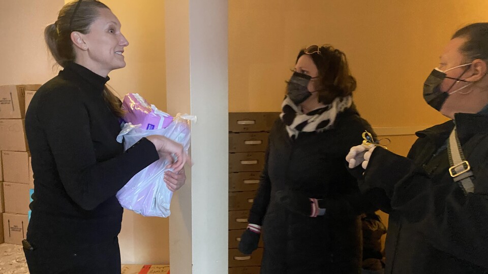 Une femme tient dans ses mains un sac de plastique. Elle discute avec deux autres femmes, dont l'une tient un ruban aux couleurs de l'Ukraine, jaune et bleu. Plusieurs boîtes sont empilées derrière elles.
