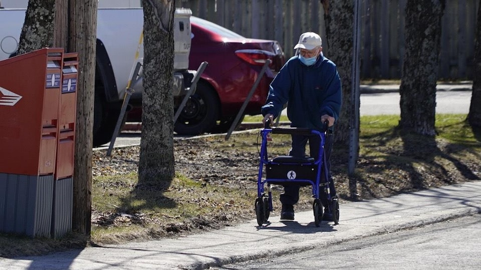 Un homme âgé marche avec un déambulateur sur un trottoir. Il porte un couvre-visage.