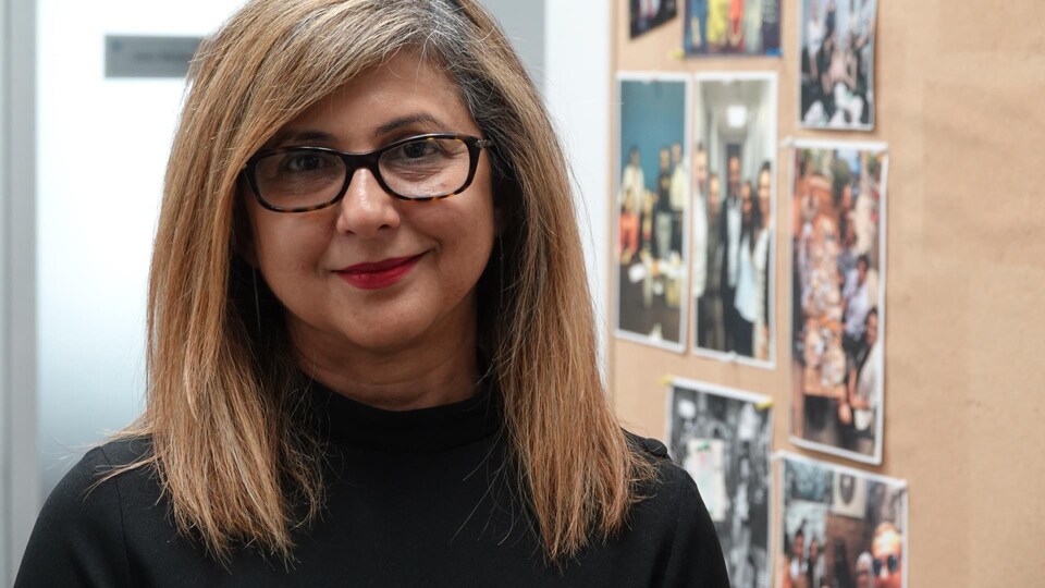 Une femme avec des lumettes pose devant un tableau où sont affichées des photos.