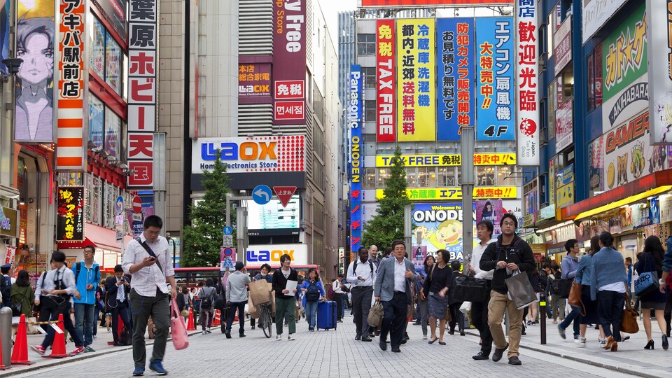 Une photo prise dans une rue du quartier Akihabara à Tokyo au Japon. On y voit des dizaines de personnes en train de marcher, dont plusieurs regardent leur téléphone. En arrière-plan et sur les côtés, on voit d'énormes panneaux colorés annonçant la présence de magasins.