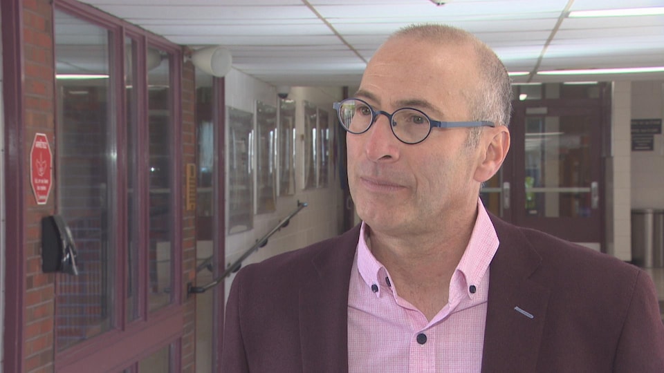 Un homme à lunettes, vêtu d'un veston rouge foncé, debout dans le hall d'entrée d'une école, l'air sérieux.