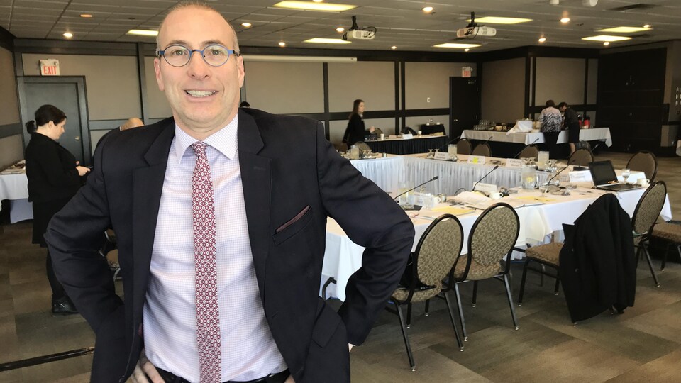 Alain Laberge se tient debout en costume-cravate dans une salle de conférence. Il est souriant avec les mains sur les hanches.