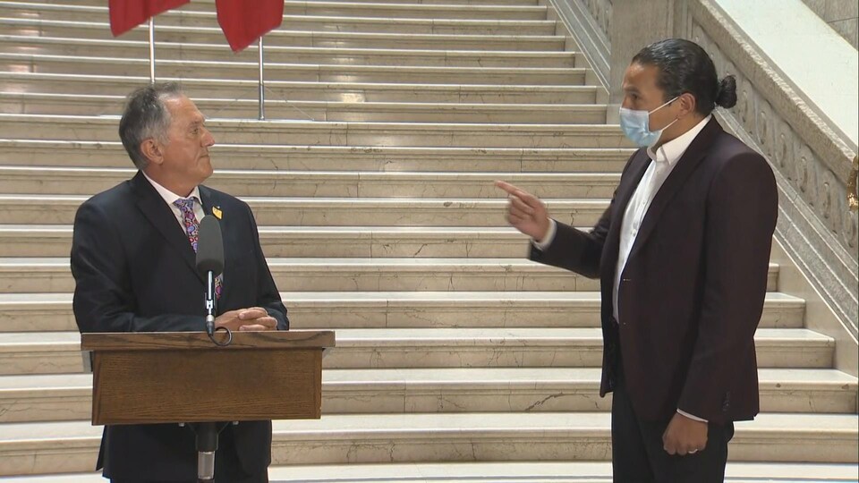 Le ministre, derrière un lutrin, regarde Wab Kinew, masqué, qui lui parle.