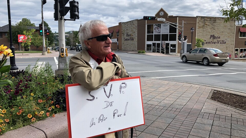 Albert Roy est debout sur un trottoir. Il porte des lunettes fumées et s'appuie sur sa pancarte, sur laquelle il a écrit « s'il vous plait, de l'air pur! »