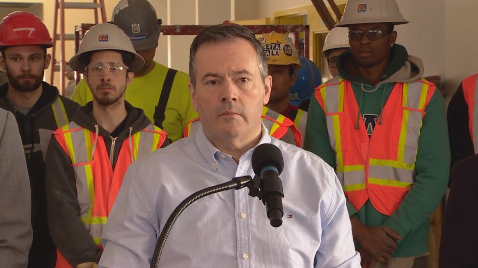 Le premier ministre albertain Jason Kenney, s'exprime devant un micro. Derrière lui, des travailleurs de la construction écoutent.