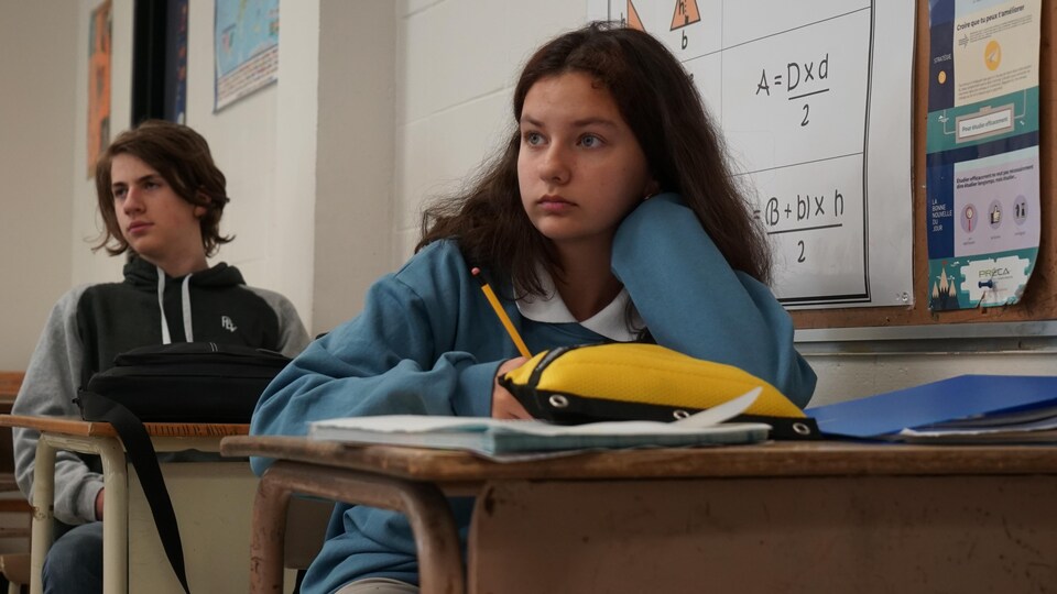Une jeune fille de 1re secondaire assise à son bureau dans sa classe regarde en avant.