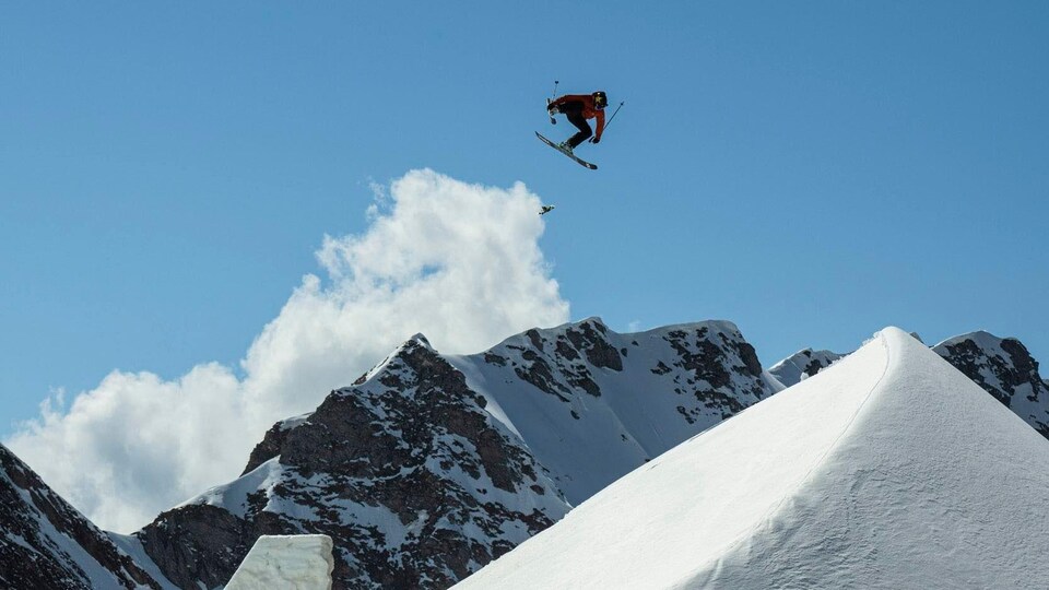 Durant un saut, il agrippe l'un de ses skis dans les airs, des montagnes derrière lui.
