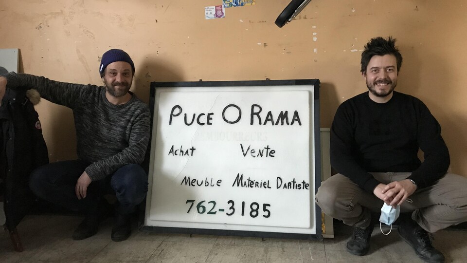 Les deux hommes assis sur le sol du magasin sourient à la caméra.
