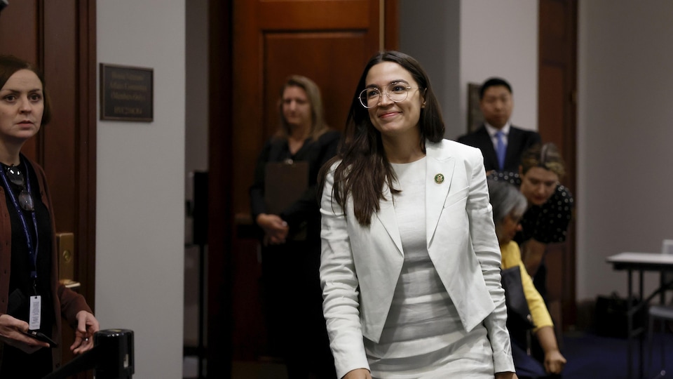 Alexandria Ocasio-Cortez marche dans un corridor sous le regard d'autres personnes.