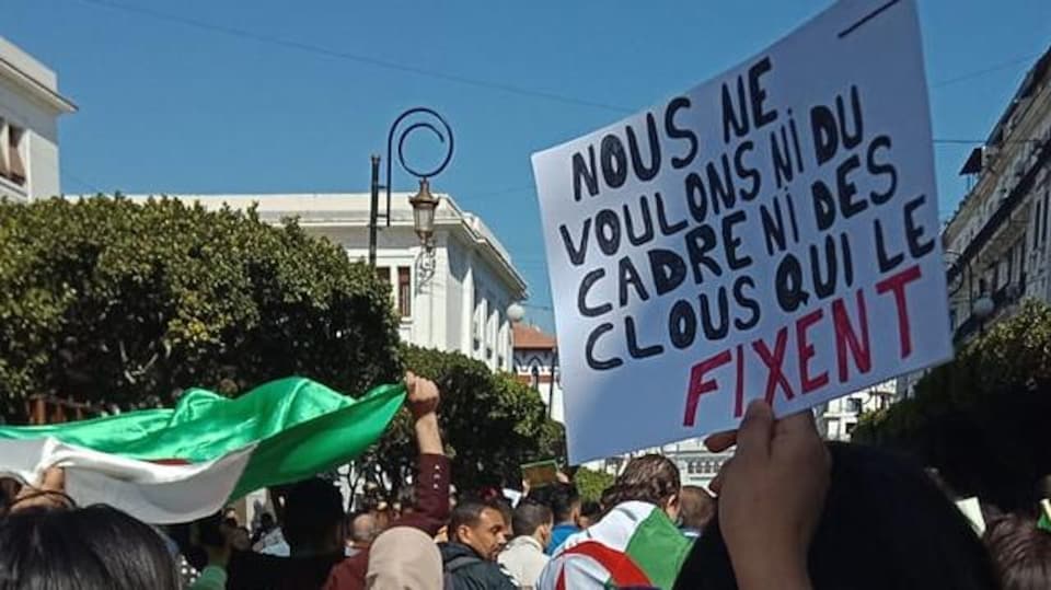 Un manifestant tient une pancarte sur laquelle on peut lire : « Nous ne voulons ni du cadre ni des clous qui le fixent ».