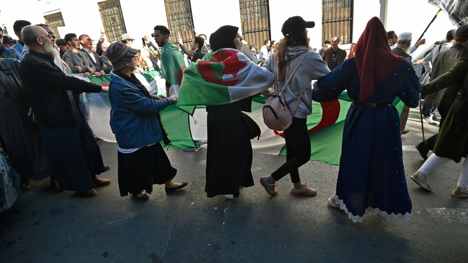 Des hommes et des femmes dansent autour du drapeau algérien en se tenant par la main.
