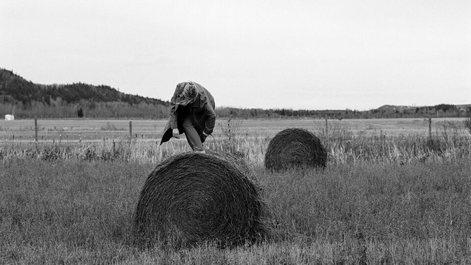 Une image noir et blanc d'Alice Cloutier-Lachance, artiste. Une jeune femme danse sur une balle de foin ronde, dans un champ à l'automne.