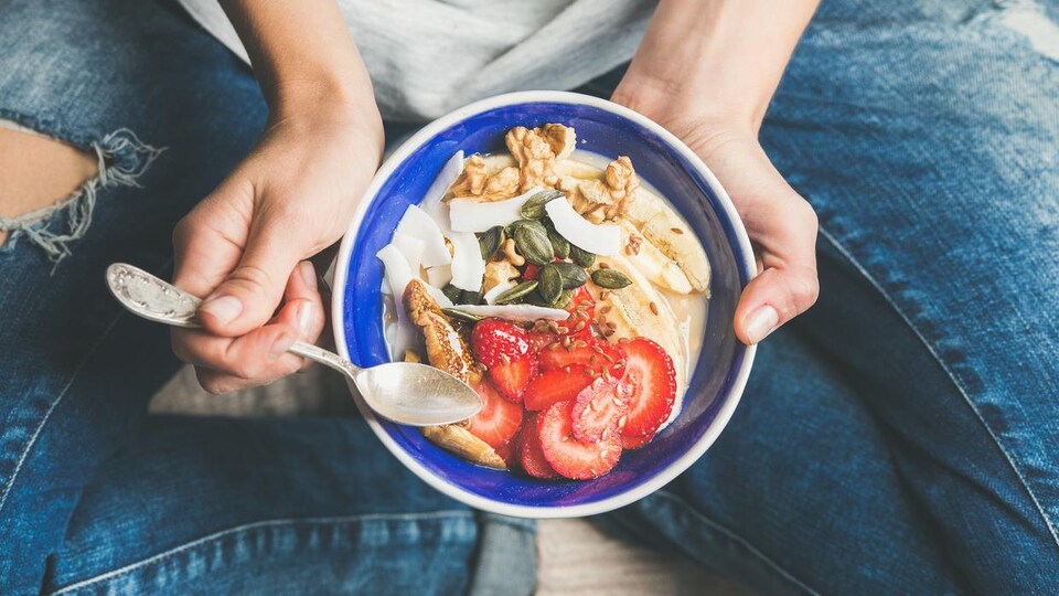 Une femme assise en tailleur tient entre ses mains un bol rempli de yaourt, fraises, graines et noix.