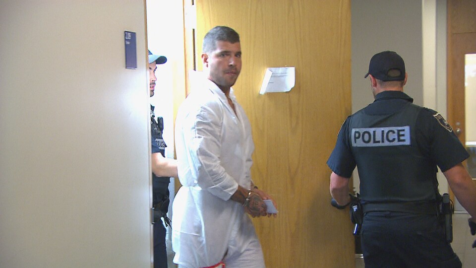 Un homme en combinaison de prisonnier est menotté et sors d'une pièce.