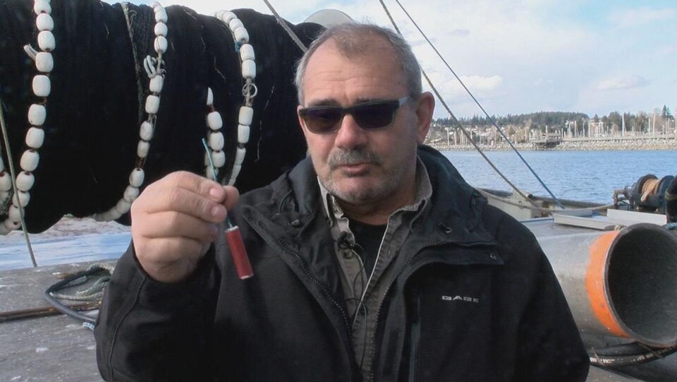 Le pêcheur se tient sur le pont de son bateau et montre un pétard de la longueur d'un doigt.