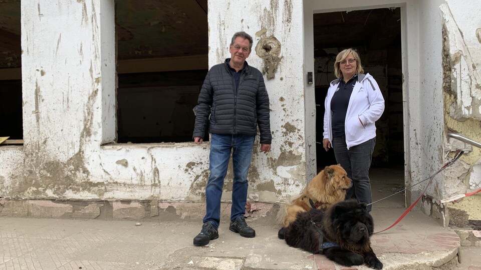 Ein Mann und eine Frau stehen mit ihren beiden Hunden vor einem baufälligen Gebäude.