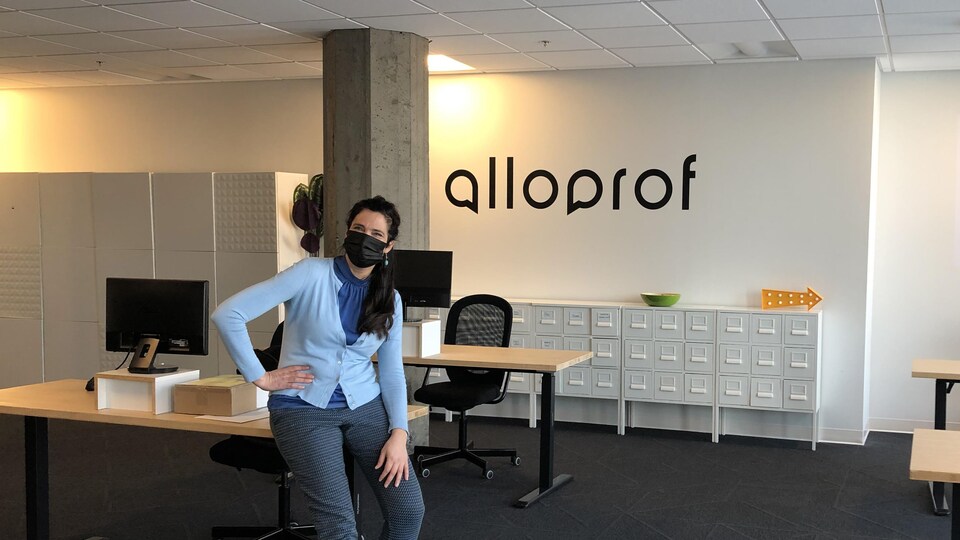 Une femme portant un masque est assise sur le coin d'un bureau. Sur le mur derrière elle est écrit : Alloprof.
