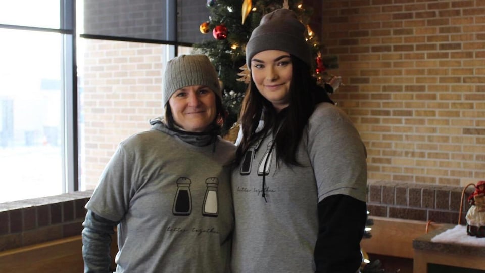 Allysa Woodrow pose avec une femme plus âgée devant un sapin de Noël. Toutes deux portent des T-shirts similaires.