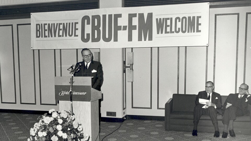 Le président au lutrin prononce un discours. Une banderole à l’arrière indique «Bienvenue CBUF-FM Welcome». 