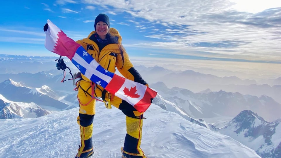 Marie-Pier Desharnais se tient au sommet d'une montagne enneigée, elle tient des drapeaux dans ses mains. 