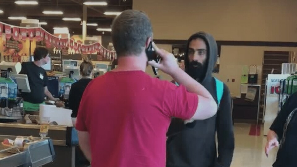 Un homme portant un chandail rouge empêche un autre homme vêtu de noir de sortir d'une épicerie.