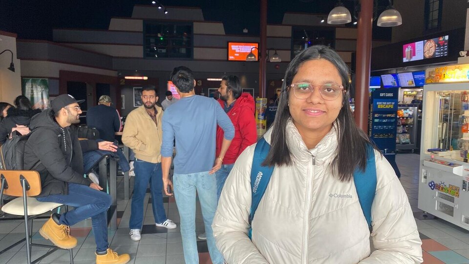 A young Indian student looks embarrassed into the entrance of a cineplex.  Behind her are several students, a slot machine, and a popcorn canteen. 