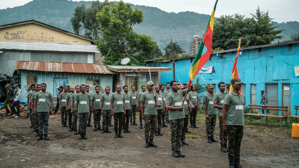 Des recrues de la milice se tiennent debout lors d'un exercice devant des bâtiments en tôle et des montagnes.