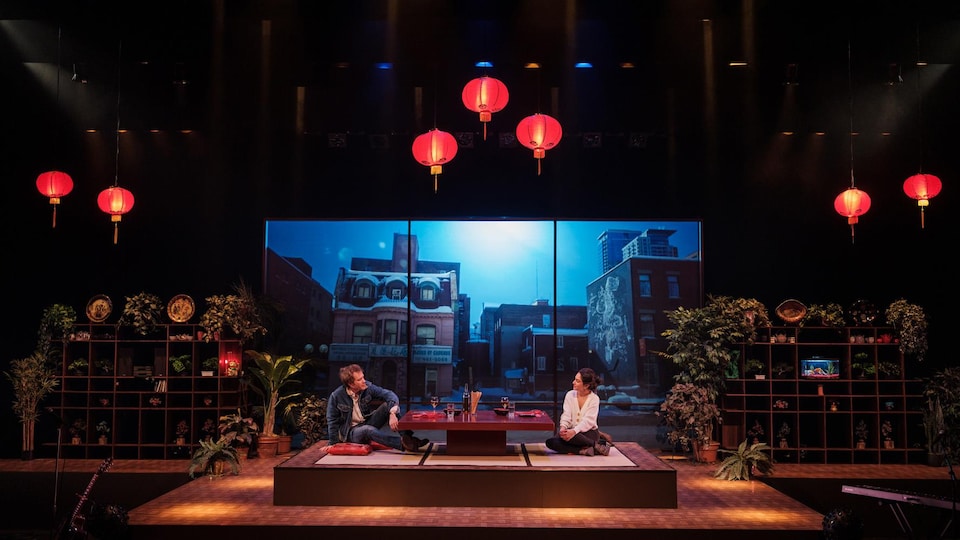 Une femme et un homme sont assis par terre de chaque côté d'une table sur une scène. Le décor évoque un restaurant asiatique : lanternes rouges, tatamis et plantes.
