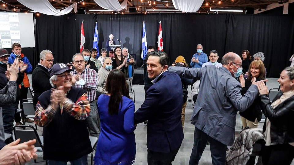 Pierre Poilievre tient la main de sa femme Anaida (à gauche) et de l'ancien maire de Trois-Rivières, Yves Lévesque (à droite), alors qu'ils avancent vers la scène, applaudis par des sympathisants. 